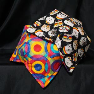 May include: Two fabric bowl cozies. One features a colorful geometric pattern with circles in red, orange, yellow, blue, and purple. The other has a black background with white cats wearing sunglasses and rainbows.
