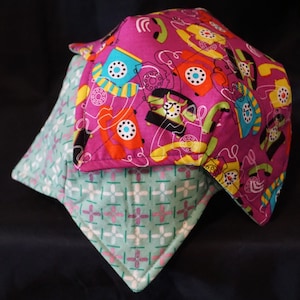 Puede incluir: Un agarrador de olla de tela rosa y verde con un patrón de teléfono retro. El agarrador de olla está hecho de dos capas de tela, con la capa superior que presenta un fondo rosa con un patrón repetido de teléfonos rotatorios coloridos y la capa inferior que presenta un patrón a cuadros verde y blanco.