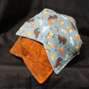 Puede incluir: Dos protectores para cuencos. El superior es azul claro con un patrón repetido de animales del bosque, como osos y zorros. El inferior es de color marrón anaranjado liso. Ambos son cuadrados con esquinas dobladas.