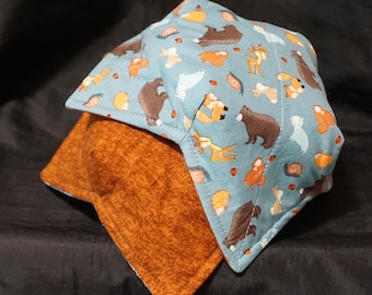May include: Two bowl cozies. The top cozy is light blue with a repeating pattern of woodland animals, including bears, foxes, and deer. The bottom cozy is a solid orange-brown color. Both are square with folded corners.