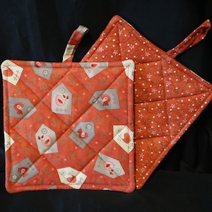 Puede incluir: Dos agarraderas de olla acolchadas rojas con un patrón floral y un diseño de casas con pájaros. Una agarradera tiene un asa de bucle.