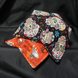 May include: A fabric bowl cosy featuring a black background with a colourful sugar skull pattern, layered over an orange fabric base printed with skulls and the text "PUT A SPELL ON YOU" in black lettering, constructed with quilted fabric.