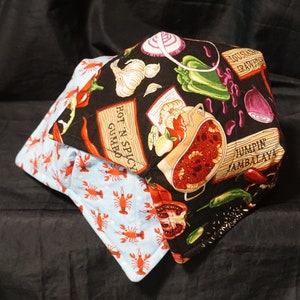 May include: A black and blue quilted pot holder with a design of various food items, including crawfish, peppers, onions, and a bowl of jambalaya. The text "Hot N Spicy Gumbo" and "Jumpin' Jambalaya" is printed on the pot holder.