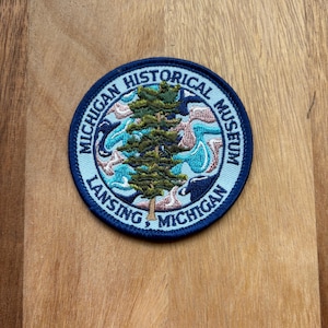 May include: Circular patch with a dark blue border featuring the text "MICHIGAN HISTORICAL MUSEUM" and "LANSING, MICHIGAN". The patch has a central design of a green tree and blue and white abstract shapes.
