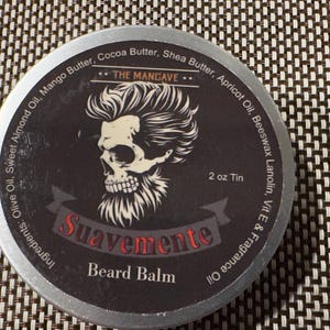 May include: A round, silver tin of beard balm. The label features a skull with a stylized hairstyle and beard. The text "Suavemente Beard Balm" is displayed in red. The tin contains 2 oz of product.