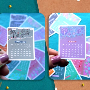 Peut inclure: Deux petits autocollants de calendrier sont pr&eacute;sent&eacute;s, l'un pour janvier et l'autre pour avril. L'autocollant de janvier pr&eacute;sente une sc&egrave;ne hivernale avec des arbres d&eacute;nud&eacute;s et le mot "janvier". L'autocollant d'avril pr&eacute;sente des arbres violets et le mot "avril".