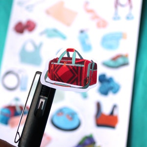 May include: A red and black duffel bag with a teal zipper and handle. The bag is on a white sheet with other stickers.