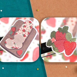 May include: Two stickers featuring strawberry designs. One sticker shows a strawberry roll cake on a wooden cutting board. The other sticker shows three strawberries with green leaves.