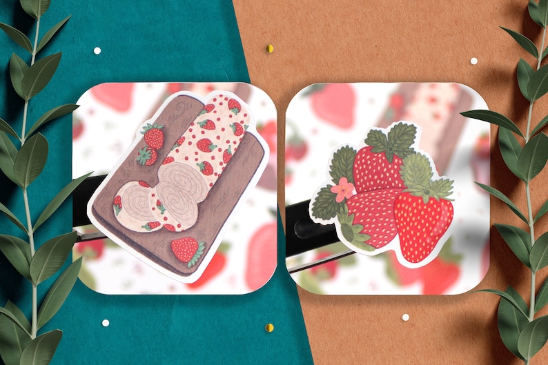 May include: Two stickers featuring strawberry designs. One sticker shows a strawberry roll cake on a wooden cutting board. The other sticker shows three strawberries with green leaves.