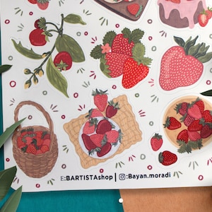 May include: A sheet of stickers featuring various illustrations of strawberries, including a basket full of strawberries, a plate of strawberries, and a strawberry plant with leaves and flowers. The stickers are on a white background with a light brown border. The text "E:BARTISTAshop | @Bayan.moradi" is printed at the bottom of the sheet.