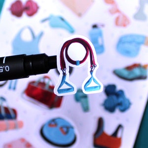 May include: A close-up of a sticker featuring a red and blue resistance band with handles. The sticker is on a white background with other fitness-themed stickers.