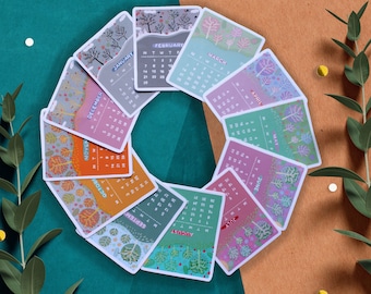 May include: A set of 12 colorful cards featuring a monthly calendar with a nature-themed design. Each card has a different color and features a different type of tree. The months of the year are printed on the cards, along with the days of the week.