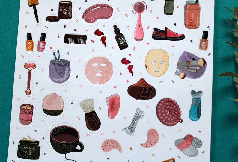 May include: A sheet of stickers featuring various beauty and self-care items, including a face mask, bath salts, a facial cleansing brush, a jade roller, a makeup brush, and a cup of tea. The stickers are on a white background with pink and purple accents.