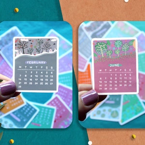 Peut inclure: Deux petites cartes de calendrier sont pr&eacute;sent&eacute;es, une pour f&eacute;vrier et une pour juin. La carte de f&eacute;vrier pr&eacute;sente une sc&egrave;ne hivernale avec des arbres d&eacute;nud&eacute;s, tandis que la carte de juin pr&eacute;sente un motif d'arbres color&eacute;s. Les cartes sont tenues sur un fond d'autres cartes de calendrier.