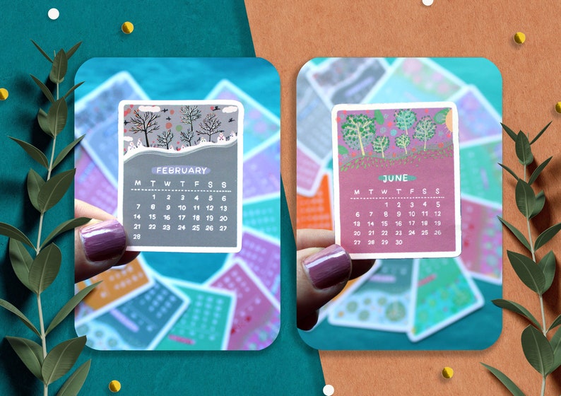 Peut inclure: Deux petites cartes de calendrier sont pr&eacute;sent&eacute;es, une pour f&eacute;vrier et une pour juin. La carte de f&eacute;vrier pr&eacute;sente une sc&egrave;ne hivernale avec des arbres d&eacute;nud&eacute;s, tandis que la carte de juin pr&eacute;sente un motif d'arbres color&eacute;s. Les cartes sont tenues sur un fond d'autres cartes de calendrier.
