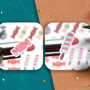 May include: Two white square stickers with a pink and white candy and a blue and white candy on a brown surface. The stickers are surrounded by green leaves.