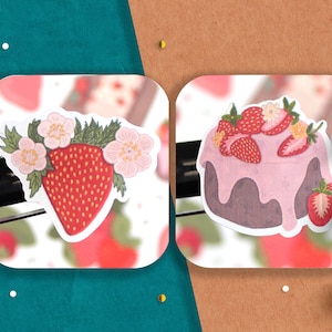 May include: Two stickers featuring strawberry designs. The first sticker shows a single strawberry with pink flowers and green leaves. The second sticker shows a strawberry cake with pink frosting and strawberries on top.