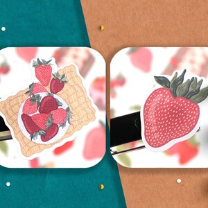 May include: Two stickers featuring illustrations of strawberries. The first sticker shows a plate of strawberries on a woven mat. The second sticker shows a single strawberry with green leaves.