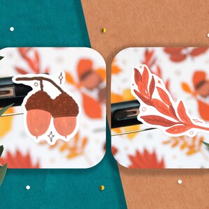 May include: Two stickers featuring autumnal designs. One sticker shows two brown acorns with pink caps. The other sticker shows a branch with red leaves.