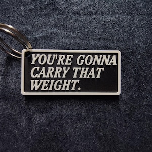 Puede incluir: Un llavero rectangular con el texto "YOU'RE GONNA CARRY THAT WEIGHT" en blanco sobre un fondo negro. El llavero tiene un borde blanco y un llavero plateado.