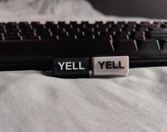 May include: Close-up of two keyboard keycaps with the word "YELL" in white. One keycap is black, the other is white. A black keyboard is in the background, resting on a light gray surface.