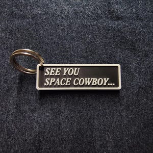May include: A rectangular black and white keychain with the text "SEE YOU SPACE COWBOY...". The keychain has a silver key ring attached. The text is in a bold, sans-serif font.