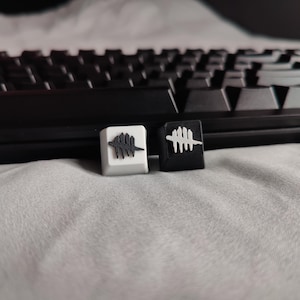 May include: Two custom keyboard keycaps, one white and one black, each featuring a stylized, dark graphic. The keycaps are in front of a black keyboard on a light gray surface. The graphic appears to be a stylized symbol.