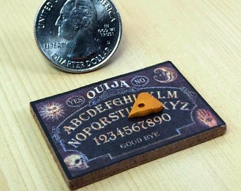 May include: A miniature Ouija board with a planchette, next to a quarter. The board is dark with white lettering and features the words "YES", "NO", the alphabet, and numbers. The planchette is orange and triangular.