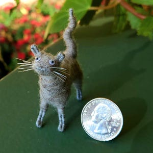 May include: A handmade miniature cat figurine made of gray yarn, with white whiskers and blue eyes. The cat stands next to a U.S. quarter coin, providing a size reference. The cat has a long tail and small ears.