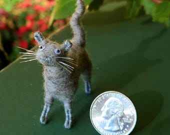May include: A handmade miniature cat figurine made of gray yarn, with white whiskers and blue eyes. The cat stands next to a U.S. quarter coin, providing a size reference. The cat has a long tail and small ears.