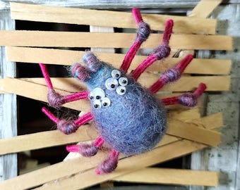 May include: A handmade, blue felt spider with multiple googly eyes and bright pink legs. The spider is positioned in front of a miniature wooden window with broken slats, creating a spooky, Halloween-themed decoration.