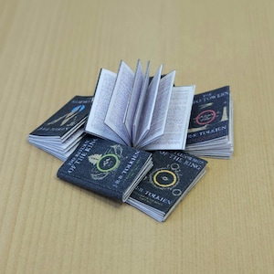 Peut inclure: Un ensemble de cinq livres miniatures reliés en carton avec des couvertures noires et des lettres dorées. Les livres sont intitulés "La Communauté de l'Anneau", "Les Deux Tours" et "Le Retour du Roi".