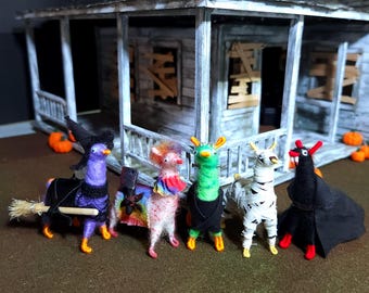 May include: A group of five Halloween-themed llama figurines. Each llama is dressed in a costume, including a witch, a colorful poncho, a chef, a mummy, and a vampire. The figurines are in front of a miniature haunted house.