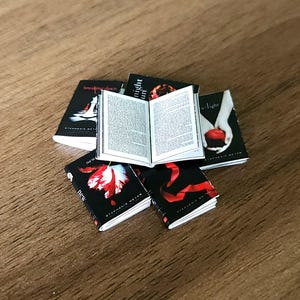 以下が含まれることがあります： トワイライト・サーガの小説をフィーチャーしたミニチュアブックセット。本は黒い表紙に赤と白のグラフィックが施されています。1冊の本が開かれており、小さな印刷されたページが表示されています。本は木製の表面に配置されています。