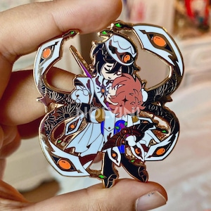 Könnte beinhalten: Detaillierter Emaille-Pin mit zwei Anime-Charakteren, die sich umarmen. Der Pin hat einen goldenen Rand mit weißen und orangefarbenen Akzenten, und die Charaktere sind in aufwendigen Outfits gekleidet. Der Pin wird von einer Hand gehalten.