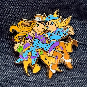 Puede incluir: Un pin de esmalte dorado con dos personajes de estilo anime. Un personaje tiene el pelo largo y rubio y lleva un sombrero, ropa morada y azul. El otro tiene el pelo rubio y lleva un atuendo azul. El pin tiene un diseño detallado.