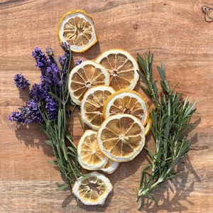 May include: A collection of dried lemon slices arranged in a vertical cluster, flanked on the left by a sprig of purple lavender flowers and on the right by a sprig of green rosemary leaves, all featuring natural textures and colors.