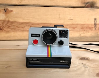 Puede incluir: Una cámara Polaroid OneStep Land Camera vintage en blanco con lente y flash negros. La cámara tiene un botón rojo, una franja de arcoíris y el texto "Polaroid Land Camera BC Series". Se adjunta una correa negra.