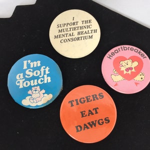 Może przedstawiać: Cztery okrągłe odznaki z różnymi wzorami. Pierwsza odznaka jest niebieska z białymi chmurami i pluszowym misiem i napisami "I'm a Soft Touch". Druga odznaka jest biała z czarnym tekstem, który brzmi "I Support the Multiethnic Mental Health Consortium". Trzecia odznaka jest różowa z pluszowym misiem trzymającym serce i napisami "Heartbreaker". Czwarta odznaka jest pomarańczowa z czarnym tekstem, który brzmi "Tigers Eat Dawgs".