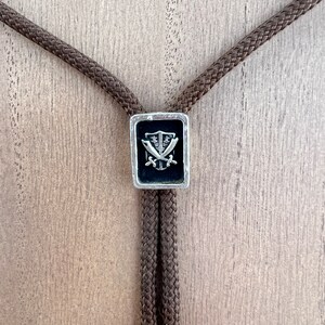 May include: A brown bolo tie with a silver rectangular pendant featuring a black shield with two crossed swords.