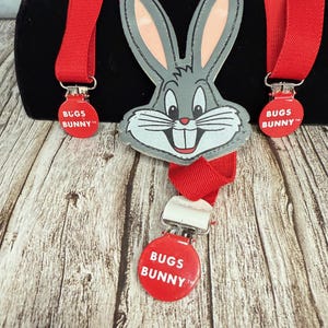 May include: Red suspenders featuring a Bugs Bunny face and three red circular clips with "BUGS BUNNY" text. The Bugs Bunny face is gray with pink inner ears and a white mouth. The suspenders are displayed on a wooden surface.