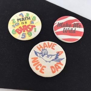 Może przedstawiać: Trzy okrągłe guziki z różnymi wzorami. Pierwszy guzik jest żółty z czarnymi cyframi i tekstem, który brzmi "Math is a Blast". Drugi guzik jest w czerwono-białe paski z tekstem "Make Me Wild". Trzeci guzik jest biały z pomarańczowym tekstem, który brzmi "Have a Nice Day" i ilustracją niebieskiego ptaka.