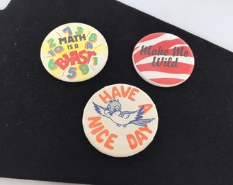 May include: Three round buttons with different designs. The first button is yellow with black numbers and text that says "Math is a Blast". The second button is red and white striped with the text "Make Me Wild". The third button is white with orange text that says "Have a Nice Day" and a blue bird illustration.