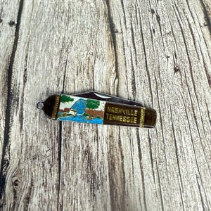 Könnte beinhalten: Ein kleines, zusammenklappbares Taschenmesser mit braunem Griff. Das Messer zeigt eine farbenfrohe Illustration einer Landschaft und die Worte "Nashville Tennessee" in goldfarbenen Buchstaben. Die Klinge ist teilweise geöffnet und zeigt eine silberne Kante.