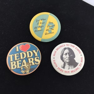 Może przedstawiać: Trzy okrągłe odznaki z różnymi wzorami. Pierwsza odznaka jest żółta i niebieska z tekstem "Just When I Knew All Life's Answers They Changed All The Questions". Druga odznaka jest niebieska z czerwonym sercem i tekstem "I Teddy Bears". Trzecia odznaka jest biała z portretem rdzennego Amerykanina i tekstem "Man Belongs To The Earth Earth Does Not Belong To Man".