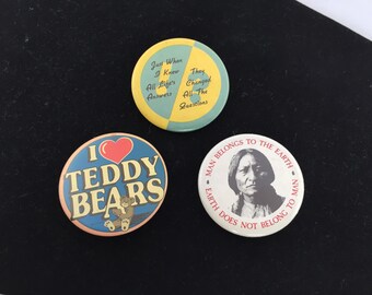 May include: Three round button pins with different designs. The first pin is yellow and blue with the text "Just When I Knew All Life's Answers They Changed All The Questions". The second pin is blue with a red heart and the text "I Teddy Bears". The third pin is white with a portrait of a Native American person and the text "Man Belongs To The Earth Earth Does Not Belong To Man".