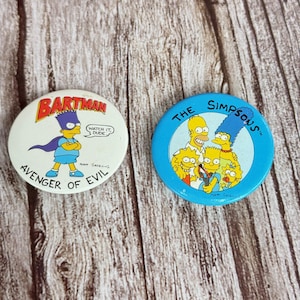 May include: Two round pinback buttons featuring characters from The Simpsons. One button has Bart Simpson as "Bartman" with the text "Avenger of Evil." The other button has the whole Simpson family with the text "The Simpsons."