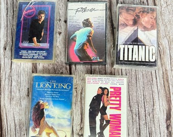 May include: Five vintage cassette tapes featuring movie soundtracks. Titles include "Cocktail," "Footloose," "Titanic," "The Lion King," and "Pretty Woman." Each tape has unique cover art with images of the films and their stars.