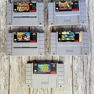 May include: Five grey Super Nintendo video game cartridges are arranged on a wooden surface. The game labels feature colourful graphics and titles such as "Nightmare", "Smac Fu", "Spider-Man", "Biometal", and "Game Master".