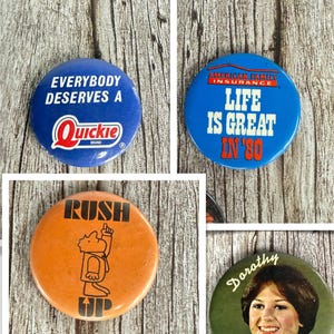 May include: A selection of vintage pin badges. One blue badge reads "EVERYBODY DESERVES A Quickie BRAND". Another blue badge says "LIFE IS GREAT IN '80" with "AMERICAN FAMILY INSURANCE". An orange badge has "RUSH UP" and a cartoon figure. A green badge has a photo of a woman named Dorothy.